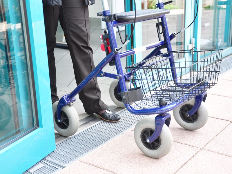 Foto: Gutjahr Eine Person mit Rollator überquert eine barrierefreie, schwellenlose Tür zu einem Gebäude mit blauen Rahmen.